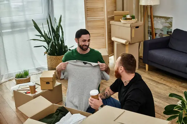 Two men, a gay couple in love, sitting on the floor of their new living room surrounded by moving boxes. — Stock Photo