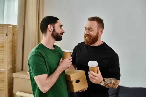 Gay couple standing side by side, holding boxes, ready to move into their new home. Boxes scattered around them. — Stock Photo