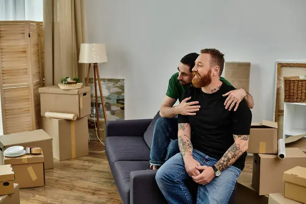 A gay couple in love sit on top of a couch in their new home, surrounded by moving boxes, embracing their new life. — Stock Photo