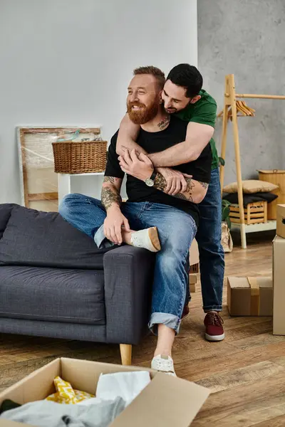 Un par de hombres se sientan encima de un sofá de felpa, abrazando su amor en medio de un movimiento caótico en su nuevo hogar. - foto de stock