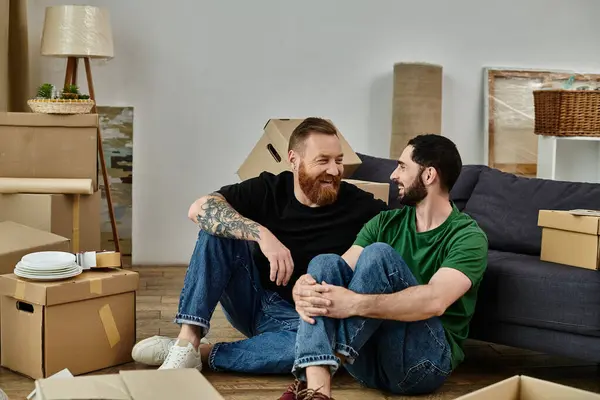 Gay couple sitting on wooden floor, surrounded by moving boxes, starting new life together in cozy home. — Stock Photo