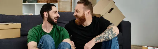 A gay couple lovingly embraces while sitting on top of a cozy couch in their new home surrounded by moving boxes. — стоковое фото