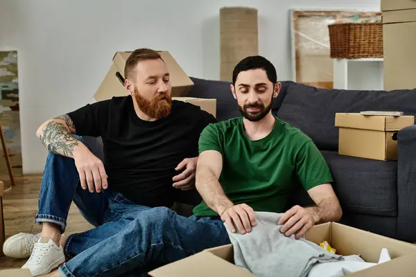 Dois homens sentam-se em cima de um sofá em sua nova casa, em meio a caixas móveis, simbolizando um novo começo em sua vida compartilhada.. — Fotografia de Stock