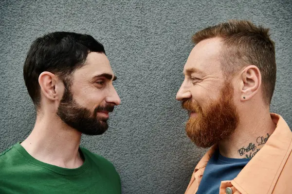 Gay couple in casual attire stand together, displaying love and affection against a grey wall backdrop. — Stock Photo
