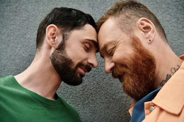 A gay couple in love, dressed in casual attire, standing closely together against a gray wall. — Stock Photo