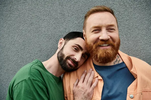 A man with a beard tenderly hugs another man in casual attire against a grey wall. — Stock Photo