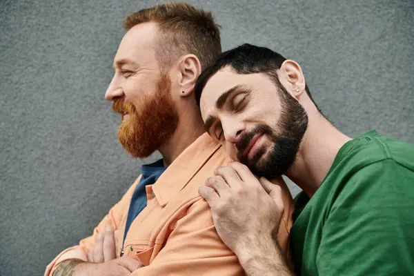 Pareja gay en atuendo casual, dos hombres con barba, se abrazan contra una pared gris en una muestra de amor y unidad. - foto de stock