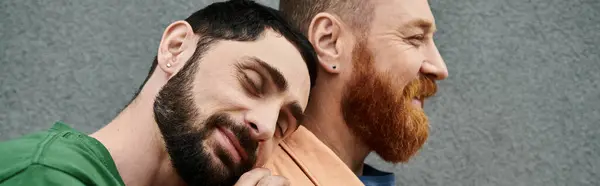 Two men with beards, a gay couple, stand side by side in casual attire against a grey wall, showing affection. — Stock Photo