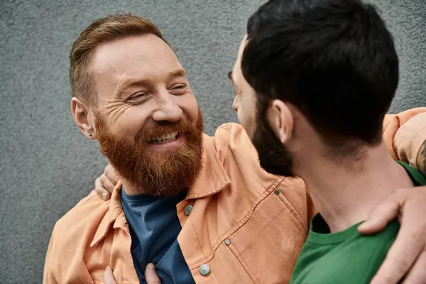 A bearded man embraces another man lovingly against a grey wall, both in casual attire. — Stock Photo