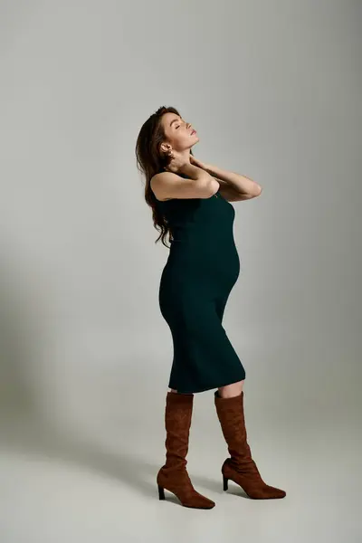 Una giovane donna incinta posa elegantemente in un abito verde e stivali marroni su uno sfondo grigio. — Foto stock