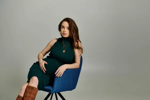 A pregnant woman in a flowing green dress gracefully sits on a vibrant blue chair. — Stock Photo