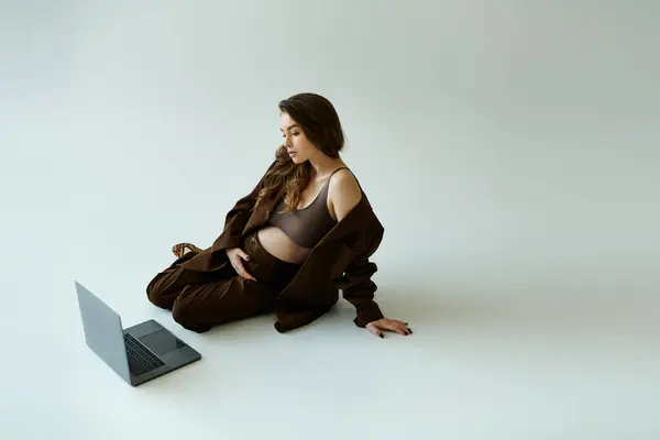 A young pregnant woman in a brown suit sits on the floor, engrossed with her laptop on a grey background. — Stock Photo
