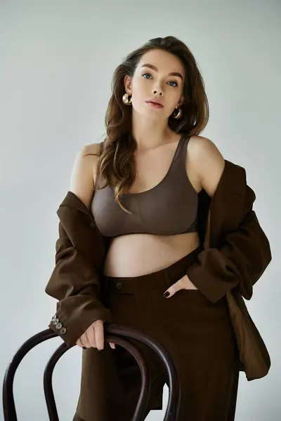 A young pregnant woman exudes grace in a brown suit, striking a pose against a grey backdrop. — Stock Photo