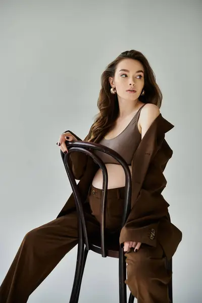 Молода вагітна жінка в коричневому костюмі сидить спокійно на стільці на сірому фоні. — стокове фото