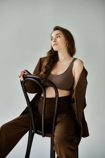 Una joven embarazada con un traje marrón se sienta en una silla sobre un fondo gris. - foto de stock