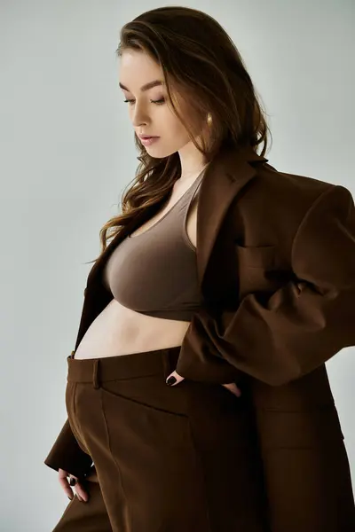 Una giovane donna incinta in un elegante abito marrone posa con fiducia su uno sfondo grigio neutro. — Foto stock