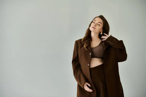 A young pregnant woman in a fashionable brown coat standing against a plain grey backdrop. — Stock Photo