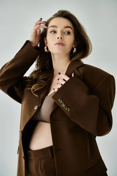 Una giovane donna incinta elegante indossa con fiducia un abito marrone con una giacca su uno sfondo grigio. — Foto stock