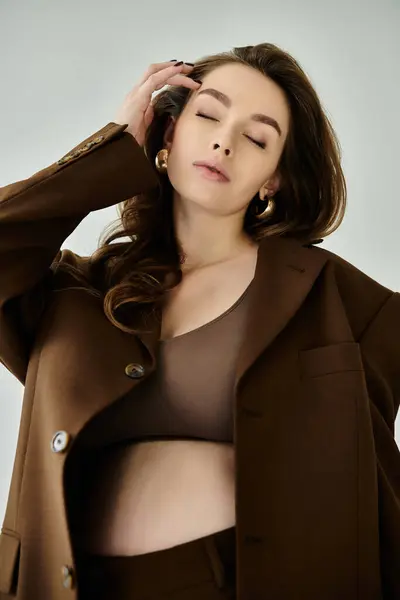 Молода вагітна жінка позує в коричневій куртці і трусиках на сірому фоні, виключаючи спокій і грацію. — стокове фото