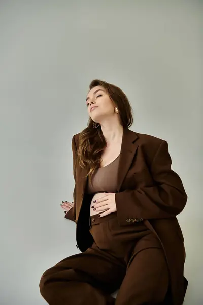 Una donna elegante e incinta in un abito marrone con una giacca, seduta con grazia su uno sgabello su uno sfondo grigio. — Foto stock
