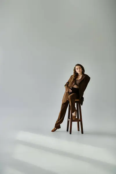 Junge schwangere Frau im braunen Anzug sitzt anmutig auf einem Holzstuhl vor grauem Hintergrund. — Stockfoto