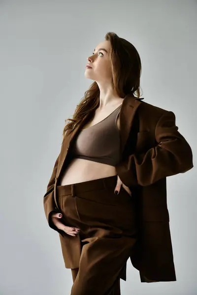 A young pregnant woman elegantly poses in a stylish brown suit with a blazer against a grey background. — Stock Photo