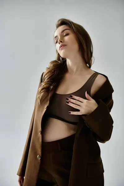 Una joven embarazada está elegantemente vestida con un traje marrón, con una chaqueta, colocada sobre un fondo gris. - foto de stock