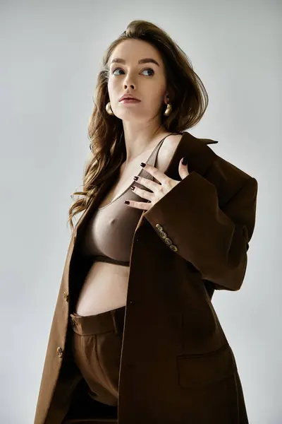 Una giovane donna incinta elegante in un abito marrone con un blazer che colpisce una posa su uno sfondo grigio. — Foto stock