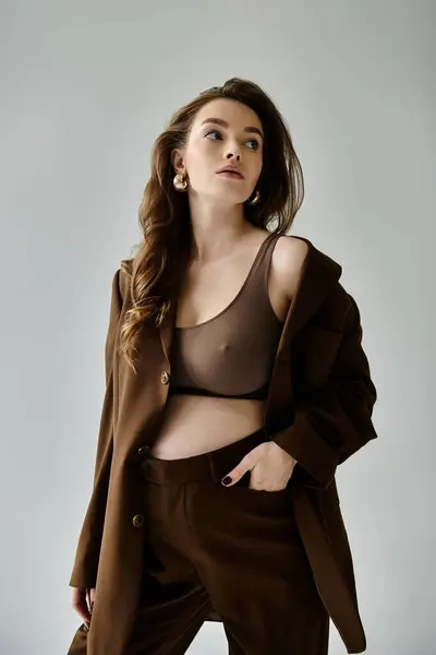 Una giovane donna incinta trasuda fiducia in un abito marrone alla moda con una giacca, colpendo una posa su uno sfondo grigio. — Foto stock