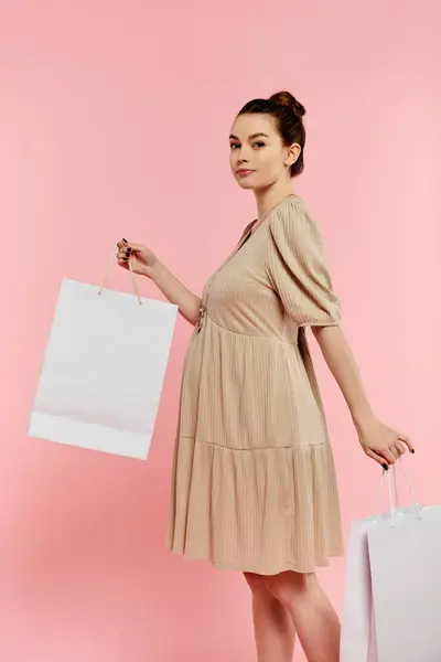 Una giovane donna incinta in un vestito tiene gioiosamente due borse della spesa su uno sfondo rosa. — Foto stock