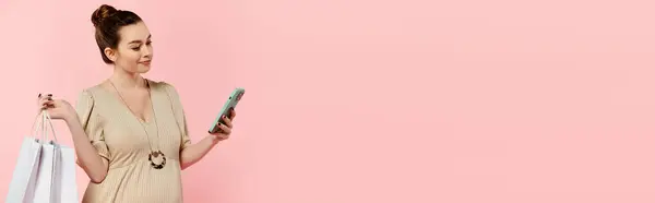 A pregnant woman in a dress holds shopping bags and a cell phone on a pink background. — Stock Photo