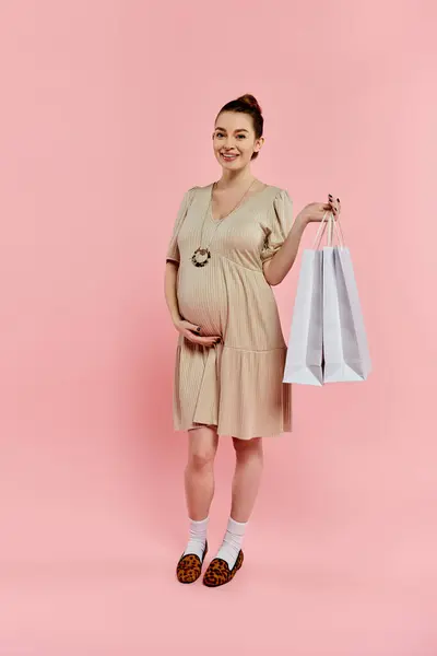 A young pregnant woman in a dress joyfully holds a shopping bag while smiling on a pink background. — Stock Photo