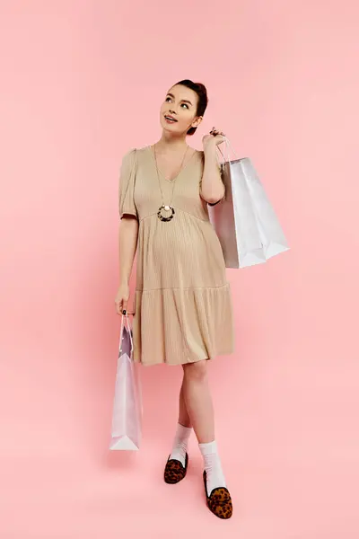 Una giovane donna incinta in un abito elegante regge con grazia le borse della spesa su uno sfondo rosa. — Foto stock