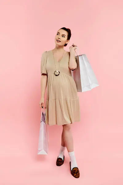 A young pregnant woman in a stylish dress gracefully holds shopping bags against a pink background. — Stock Photo