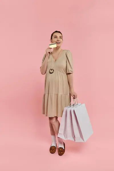 Uma jovem mulher grávida elegante em um vestido equilibrando dois sacos de compras e um cartão de crédito em um fundo rosa. — Fotografia de Stock