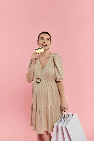 Uma jovem grávida alegremente segura sacos de compras e um cartão de crédito em um fundo rosa. — Fotografia de Stock