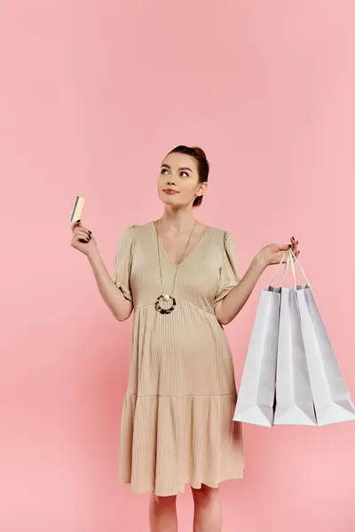 A stylish pregnant woman in a dress balancing two shopping bags and a credit card against a pink background. — Stock Photo
