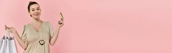 A pregnant woman in a dress, holding a credit card and shopping bags on a pink background. — Stock Photo