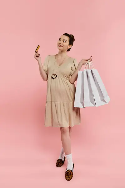A pregnant woman in a dress holds two bags and a credit card against a vibrant pink background. — Stock Photo