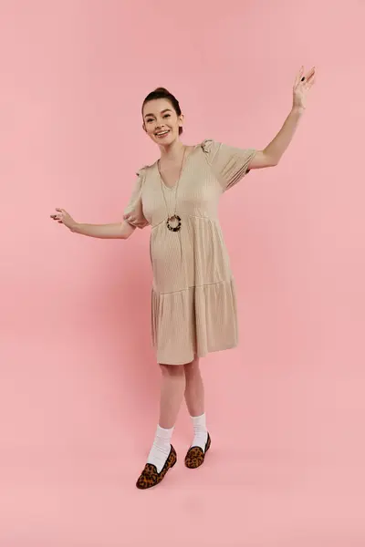 A young pregnant woman in a beige dress strikes a graceful pose on a pink background. — Stock Photo