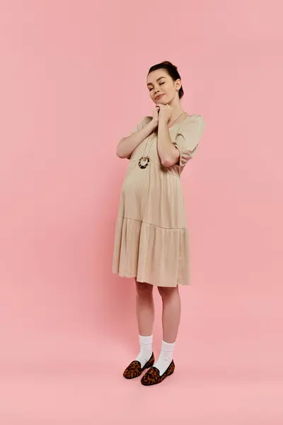 A young pregnant woman gracefully poses in a beige dress against a pink background. — Stock Photo