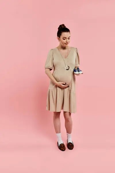 A pregnant woman in a dress lovingly holds a small baby shoe against a soft pink background. — Stock Photo