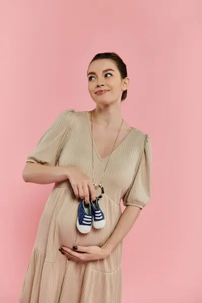 A young pregnant woman in a flowing dress gently holds a pair of baby shoes on a vibrant pink background. — Stock Photo