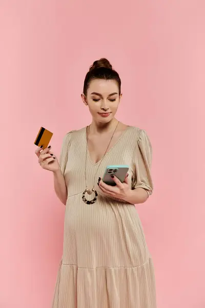 A young pregnant woman in a dress holding a cell phone and a credit card against a pink background. — Stock Photo