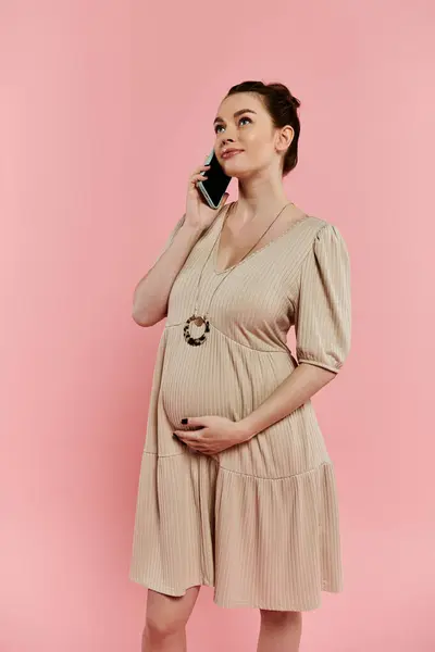 A stylish pregnant woman in a dress chatting on a cell phone against a vibrant pink backdrop. — Stock Photo