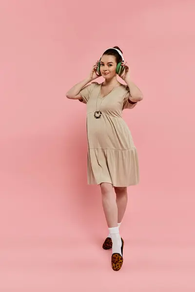 A pregnant woman gracefully stands wearing a dress, listening to music through headphones on a vibrant pink background. — Stock Photo