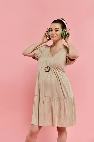 Una mujer embarazada con estilo en un vestido que fluye escucha música a través de auriculares contra un vibrante telón de fondo rosa. - foto de stock