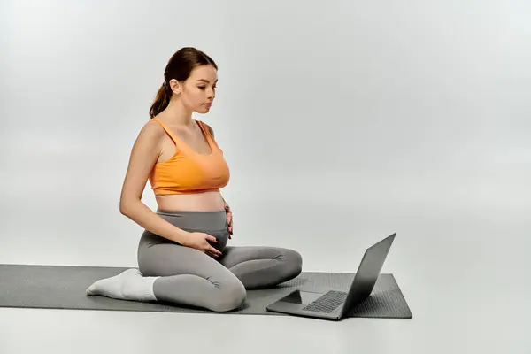 Eine sportliche Schwangere sitzt auf einer Yogamatte, Multitasking mit Laptop. — Stockfoto