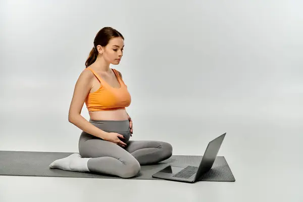 A sporty pregnant woman sits on a yoga mat, multitasking with a laptop. — Stock Photo