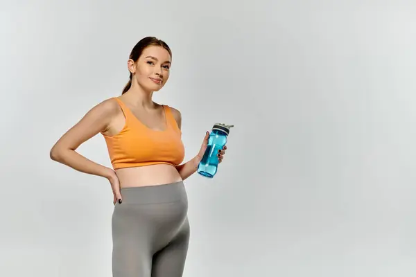 Eine junge und sportliche Schwangere in aktiver Kleidung posiert selbstbewusst mit einer Wasserflasche vor grauem Hintergrund. — Stockfoto